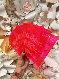 a person holding up three pink menus in front of white paper flowers and petals