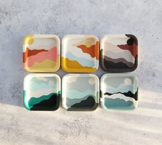 four square dishes with mountains painted on them in different shades of blue, pink, yellow and green