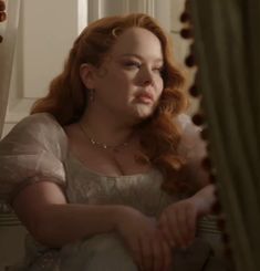 a woman with long red hair sitting in front of a window wearing a white dress