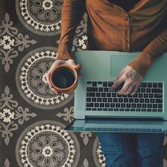 a person holding a coffee cup and using a laptop computer on a patterned tablecloth