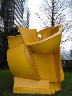 a large yellow sculpture sitting on top of a lush green field next to tall buildings