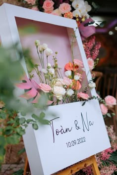 a sign with flowers on it sitting in front of a bunch of flowers and greenery