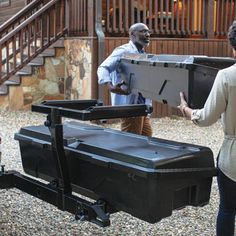 two people unloading a large black box onto a dolly cart in front of a house