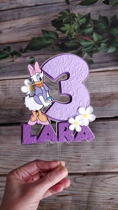 a hand holding up a purple number three shaped like a duck with flowers in front of it