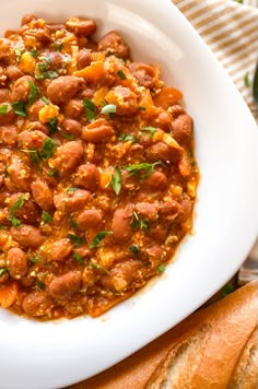 a white bowl filled with beans and bread on top of a wooden table next to a baguette