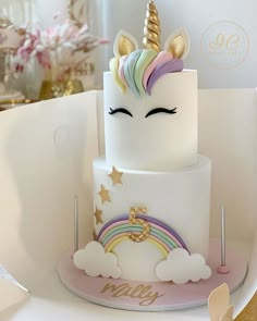 a three tiered cake with a unicorn face and rainbow horn on top, sitting on a table