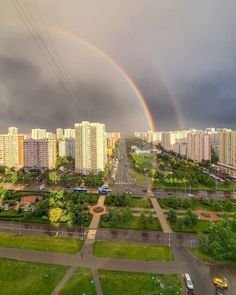 two rainbows in the sky over a city with tall buildings and green grass on both sides