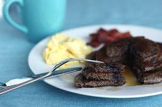 a white plate topped with pancakes and bacon next to a cup of coffee on a blue table cloth