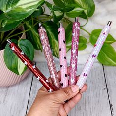 a hand holding five pink and purple glittered pens next to a potted plant