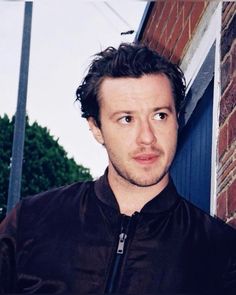 a man standing in front of a brick building wearing a black jacket and looking at the camera