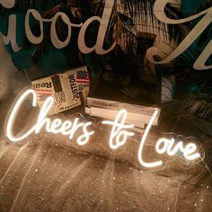 there is a neon sign that says cheers to love next to books on a bed