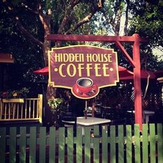 a wooden sign that says hidden house coffee in front of a green fence and trees