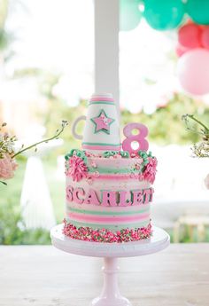 a pink and green birthday cake with stars on top is sitting on a table in front of balloons