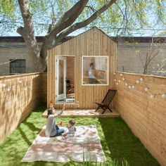 a woman and child sitting on the grass in front of a small wooden building with windows