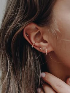 a woman wearing gold ear piercings and a white top is looking down at her left ear