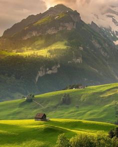 a house in the middle of a lush green field with mountains in the background at sunset