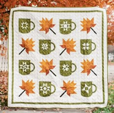 a quilted table runner with coffee mugs and autumn leaves on it in front of a tree