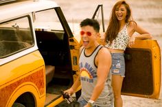 a man carrying a suitcase next to a woman on the beach with a yellow truck behind him