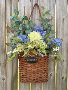 a wicker basket with blue and white flowers hanging on a wooden fence, next to a welcome sign