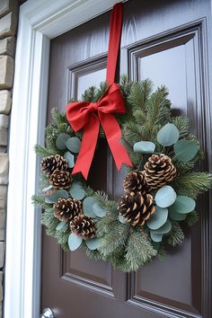 a wreath with pine cones and red ribbon hanging on a door