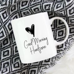 a white coffee mug with the words good morning handsome on it sitting on a black and white blanket