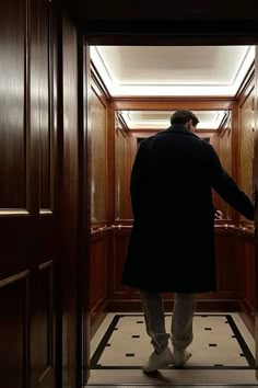 a man in a black coat and white pants is walking into a dark room with wood paneling