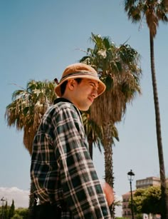 a man in plaid shirt and hat standing next to palm trees with his skateboard