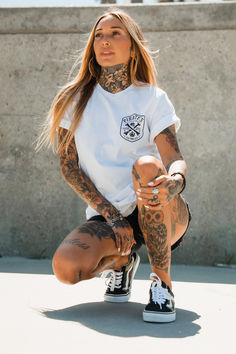 a woman with tattoos squatting down on the ground while holding a skateboard in her hands