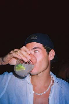 a man in a blue shirt drinking from a drink glass while wearing a baseball cap