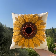 a hand holding up a decorative pillow with a sunflower on it