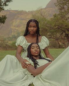 two women sitting on the ground with trees in the background, one wearing a white dress