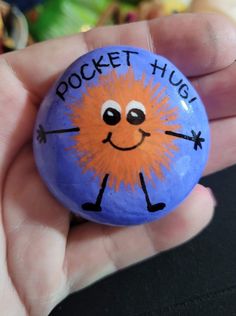 a hand holding a blue and orange rock with a drawing on it that says pocket hug