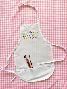 an apron with writing on it and two pencils in the pocket, sitting on a pink gingham tablecloth