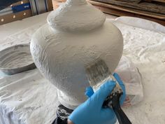 a person in blue gloves holding a paintbrush next to a white vase on a table