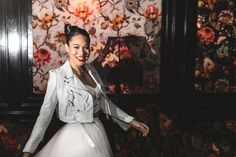 a woman standing in front of a wall with flowers on it and wearing a white dress