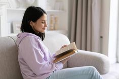 a woman sitting on a couch holding a book and looking at the pages in front of her