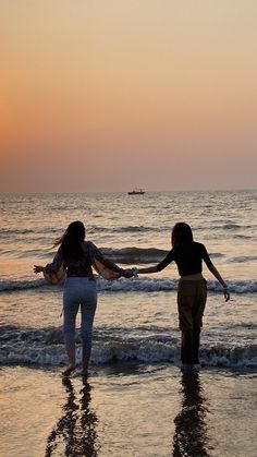 two women are walking on the beach holding hands and looking at the water as the sun sets