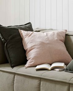 a couch with two pillows and an open book on it's back end, in front of a white wall