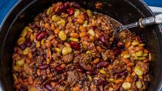 a pot filled with beans and meat on top of a blue table cloth next to a silver spoon