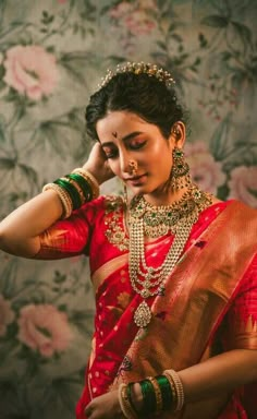 a woman in a red and gold sari with jewelry on her neck, standing against a floral wallpaper