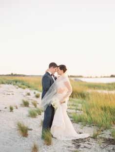 a bride and groom standing in the sand at sunset on their wedding day, posing for a photo