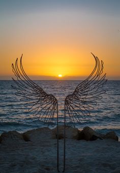 the sun is setting over the ocean with an angel's wing sculpture in front of it