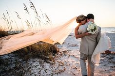 a bride and groom hug on the beach with their wedding veil blowing in the wind