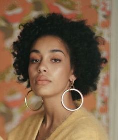 a woman wearing large hoop earrings and a yellow sweater looks off into the distance while standing in front of a floral wall