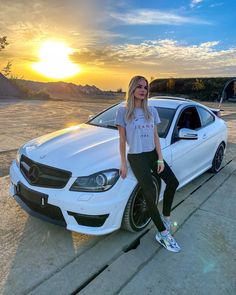 a woman standing next to a white car with the sun setting in the back ground