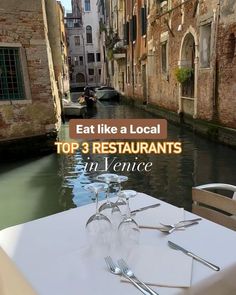 an empty table with two wine glasses and silverware on it in front of a canal