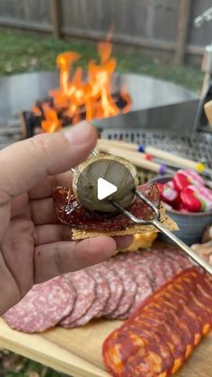 a person is holding a knife over some meats on a cutting board with flames in the background