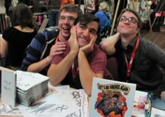 three men are sitting at a table with books and cds on it, smiling for the camera