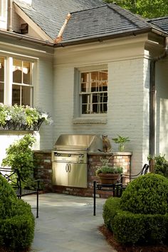 an outdoor kitchen is shown in front of a white house with bushes and shrubs around it