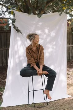 a woman sitting on top of a stool in front of a white sheeted backdrop